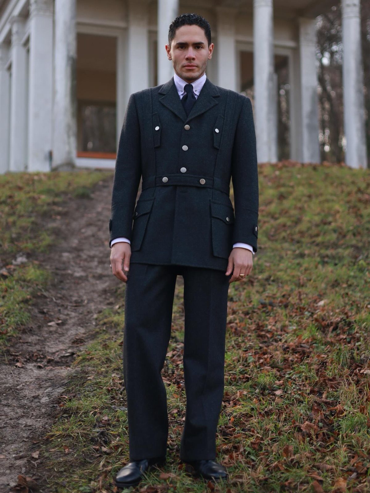 Man wearing a dark charcoal grey vintage 1880s style suit standing outdoors.