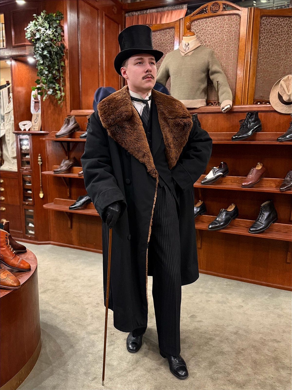 Man in 1880s attire wearing a top hat and fur-collared overcoat in a shop.