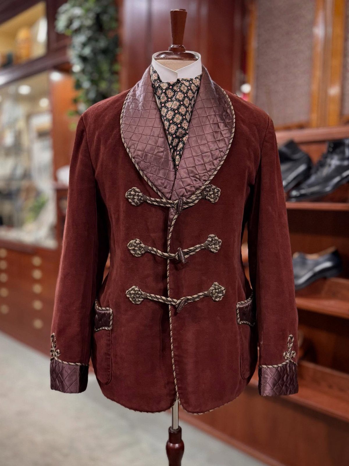 Maroon velvet smoking jacket with quilted shawl collar and gold braided fasteners on mannequin.