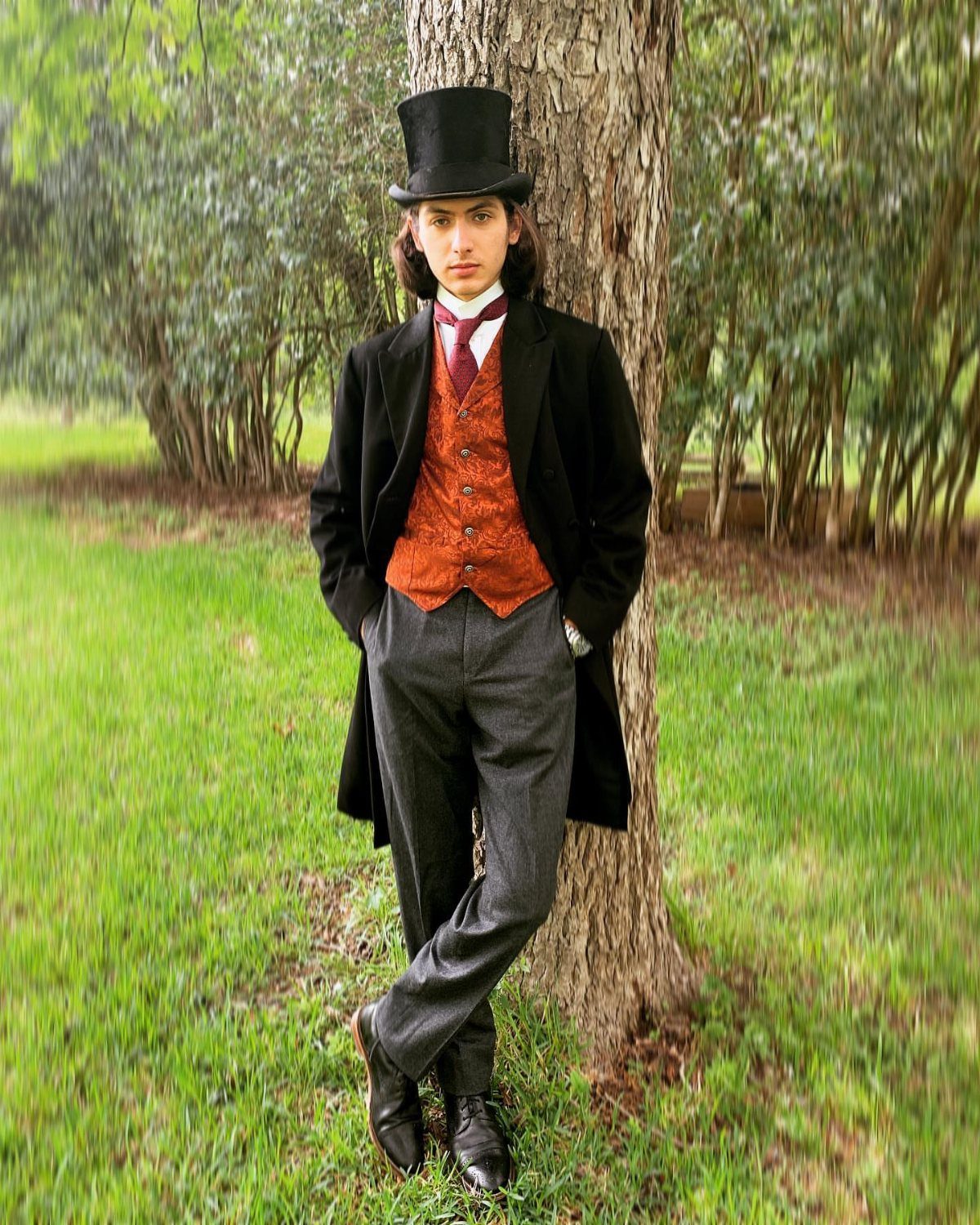 Man in 1880s attire with top hat and frock coat leaning against a tree.