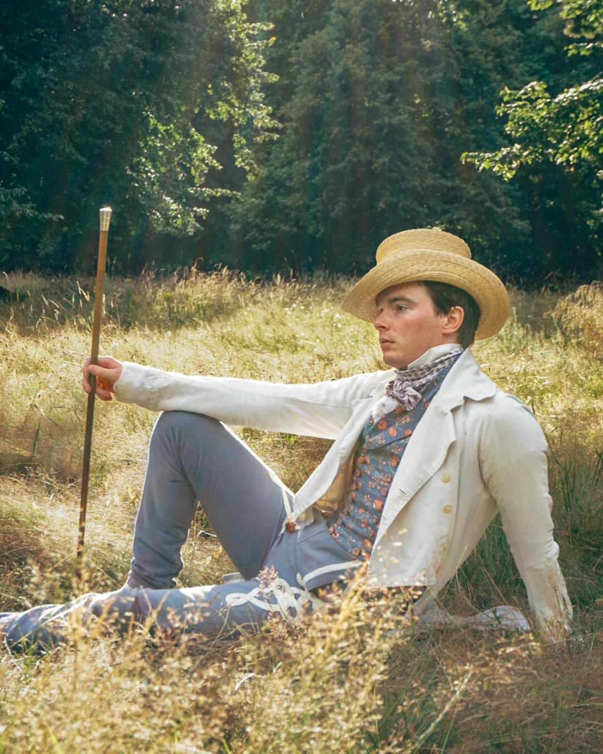 Man in 18th century men's fashion reclining in a grassy field with a hat.