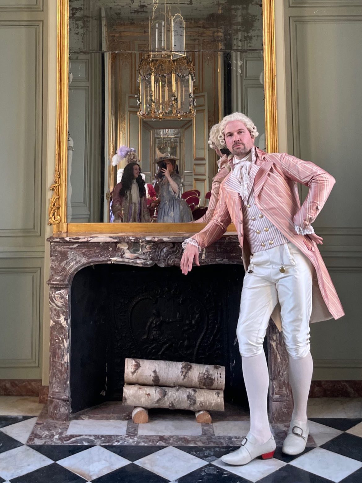 Man in pink striped 18th century frock coat and powdered wig by a fireplace.