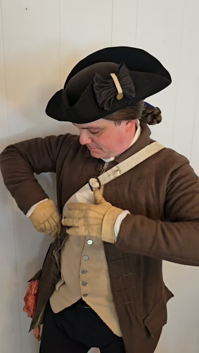 Man in 18th century men's fashion wearing a brown coat and tricorne hat.