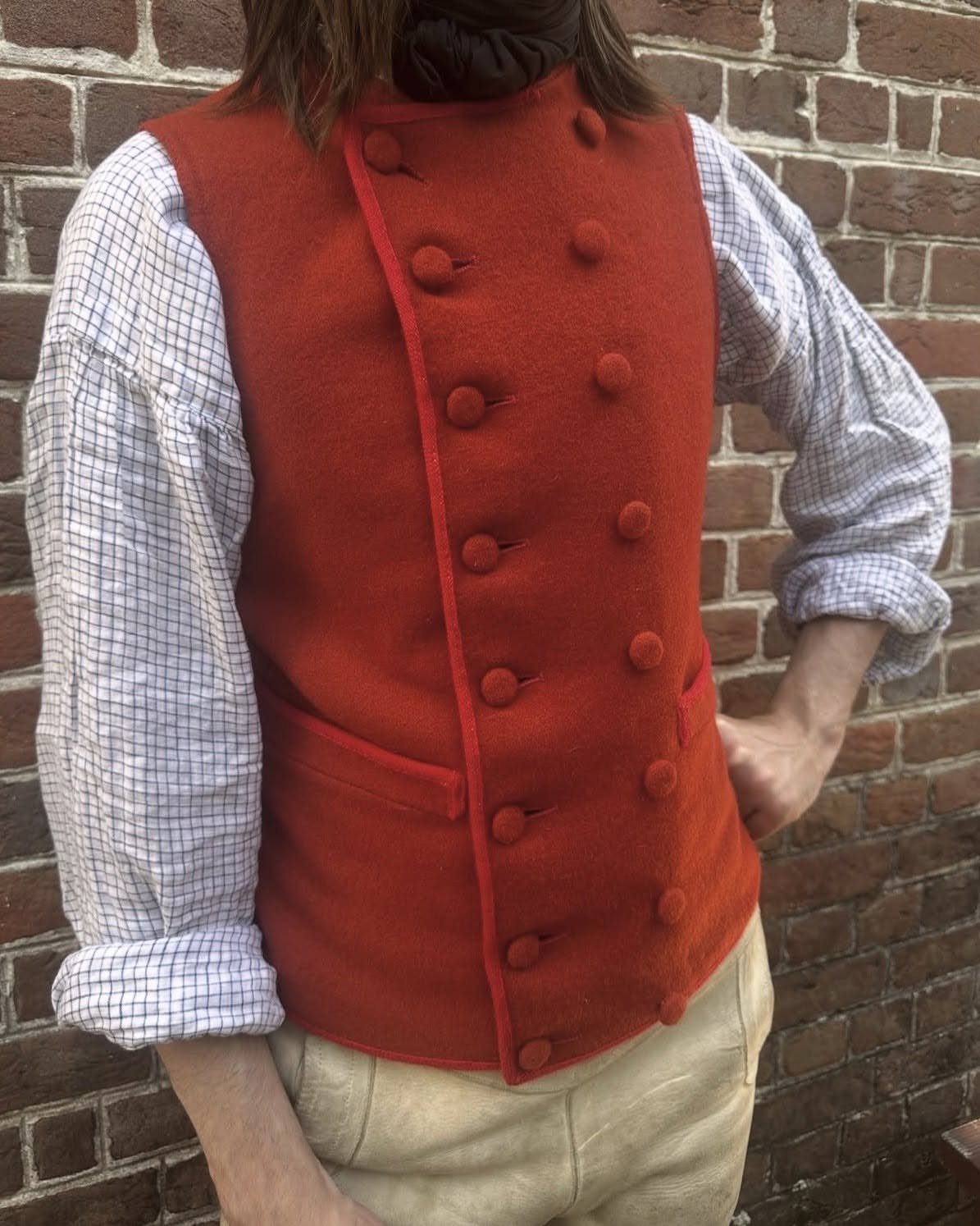 Man wearing a red 18th century waistcoat and checkered shirt against brick wall