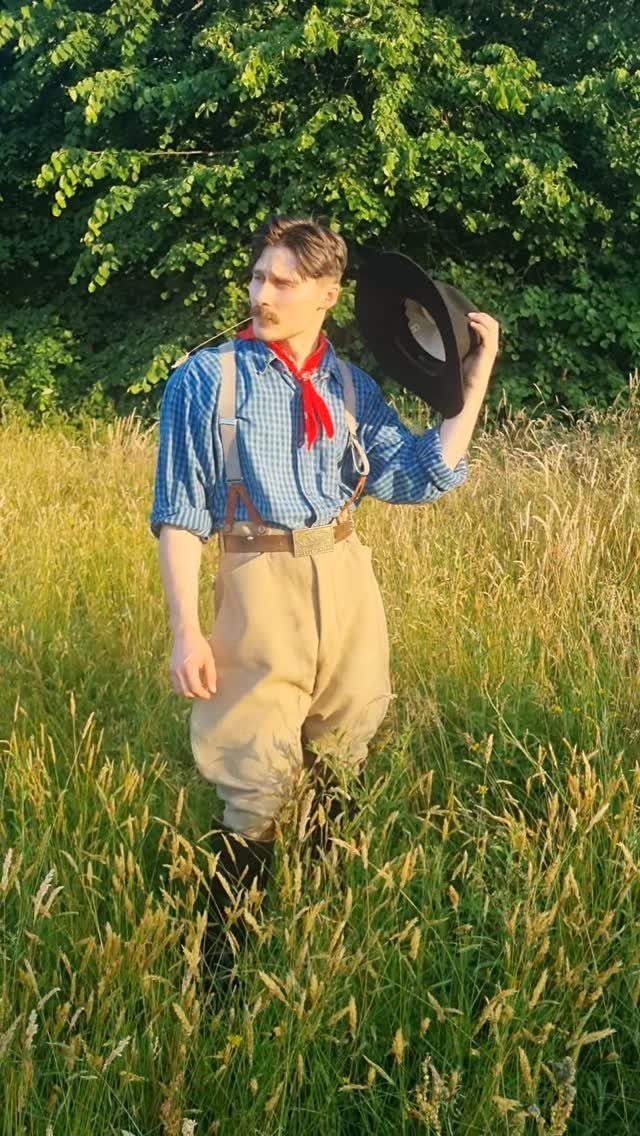 Man in 1920s working-class attire standing in a sunlit grassy field with trees.