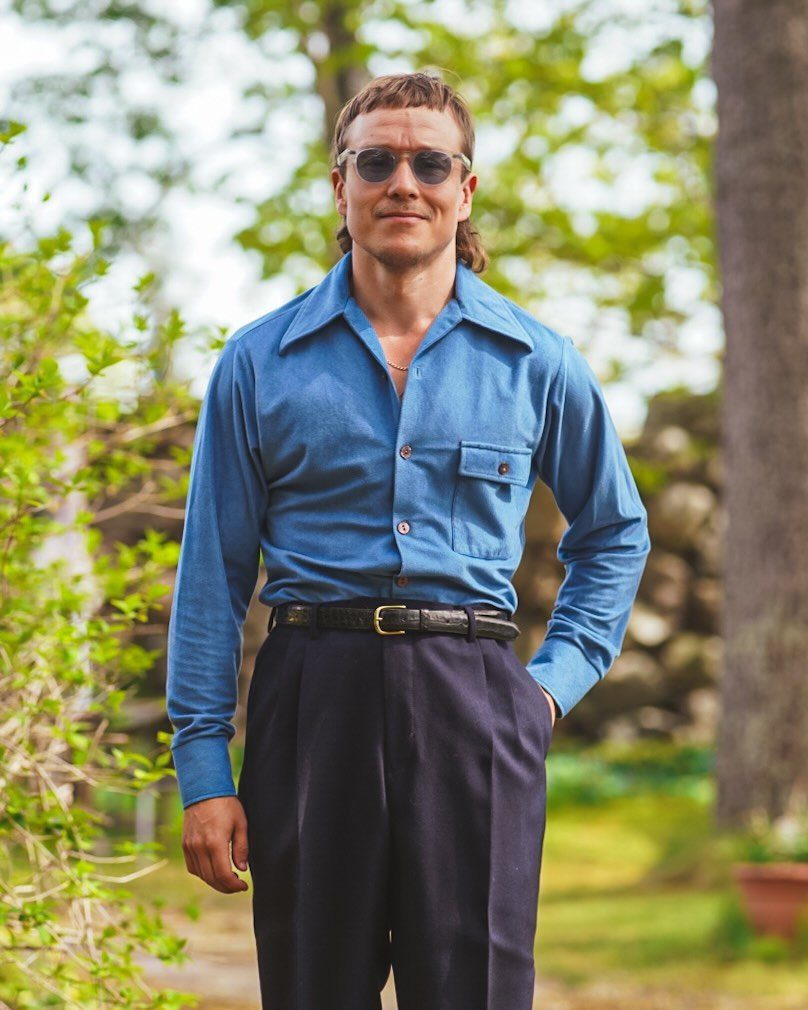 Man wearing a wide-collared blue shirt and high-waisted black trousers in a garden.