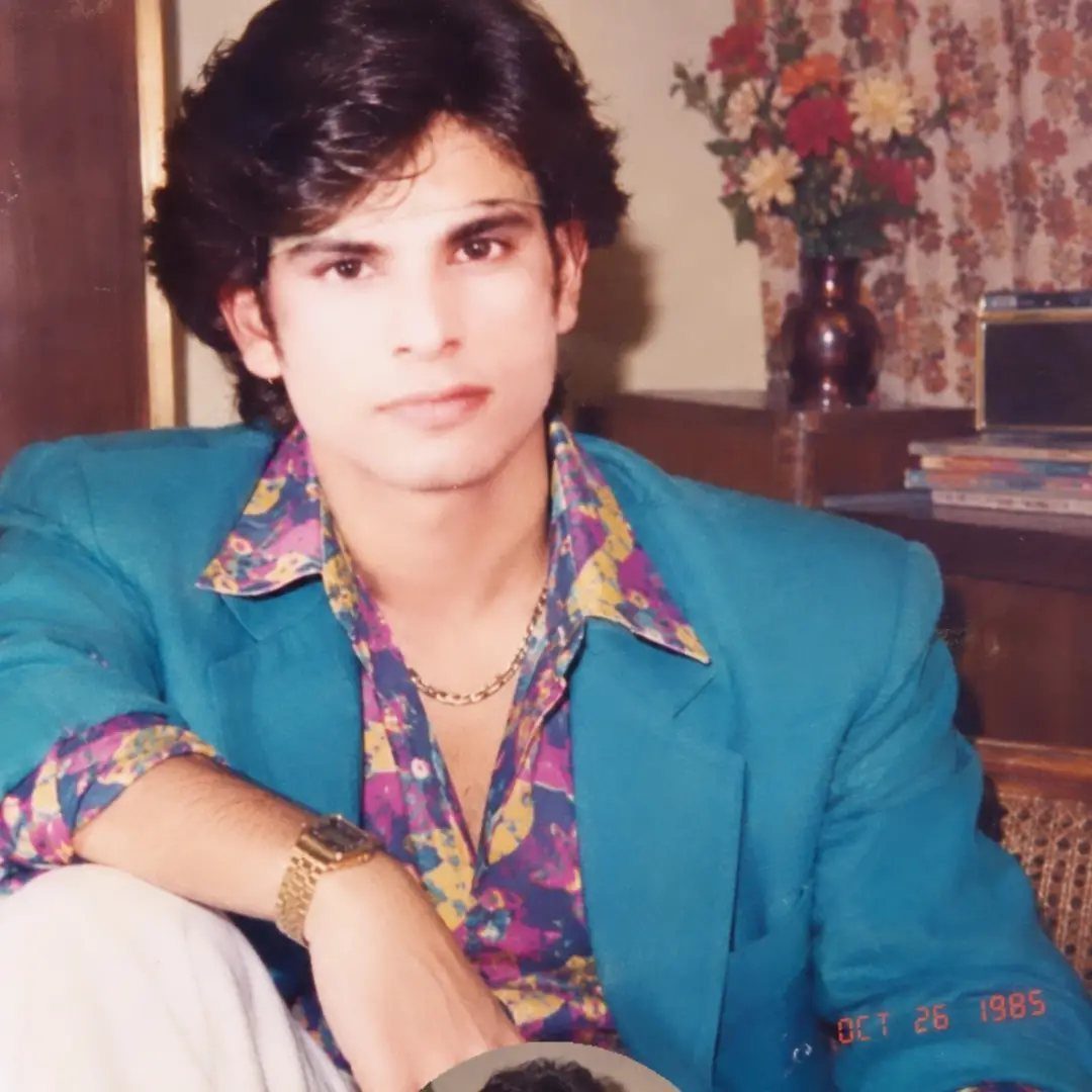 Man in a bright turquoise blazer and colorful floral print shirt posing indoors.