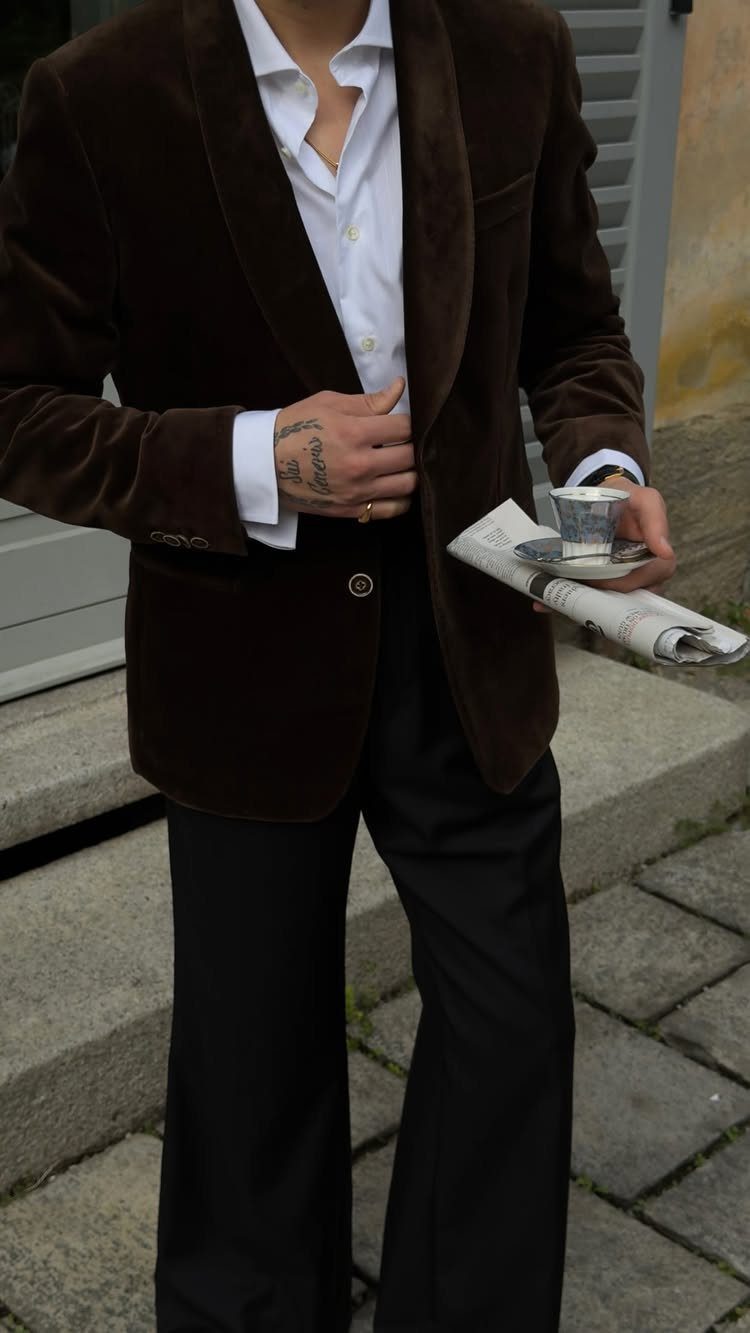Man in brown velvet blazer and white shirt holding espresso and newspaper.