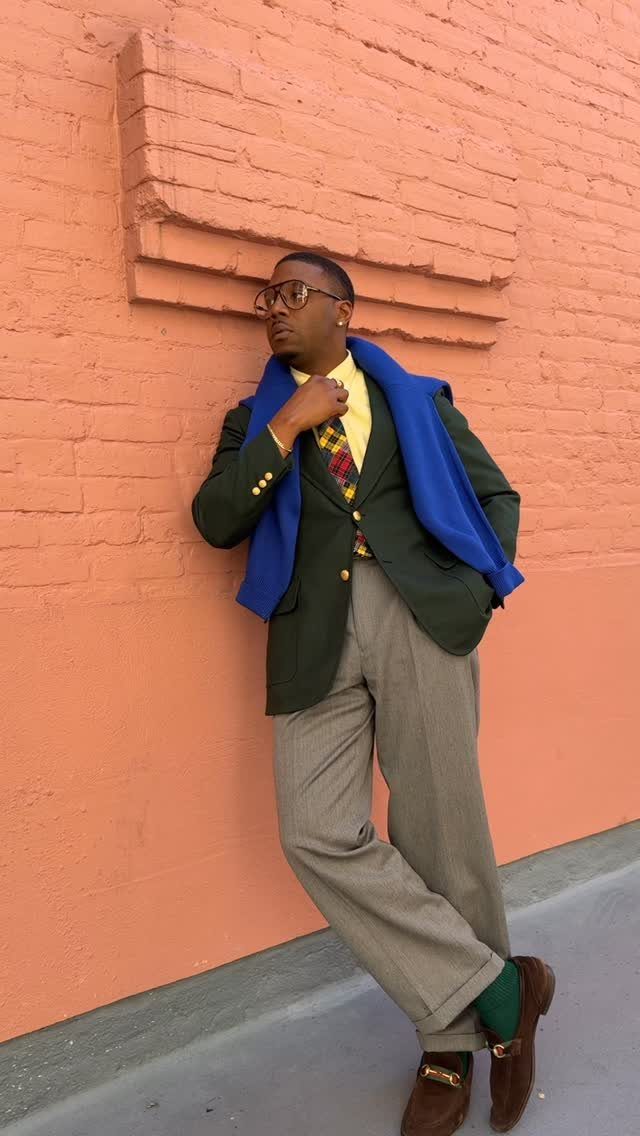 Man in a green blazer and yellow shirt leaning against a red brick wall.