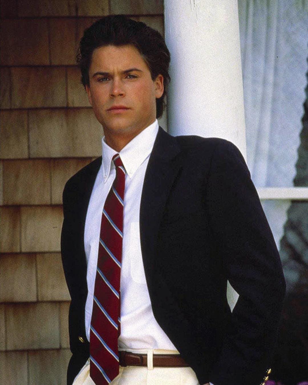 Man wearing a navy blazer, striped tie, and khaki pants leaning against a pillar.