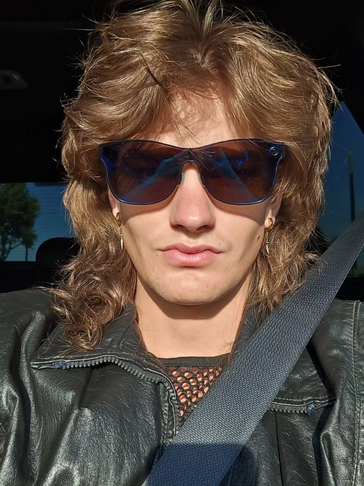 Man with mullet wearing a black leather jacket and dark sunglasses in a car.