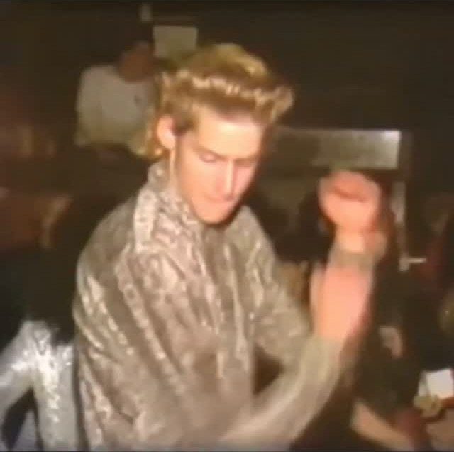 Man with blonde hair wearing a shimmering silver metallic shirt in a dim room.