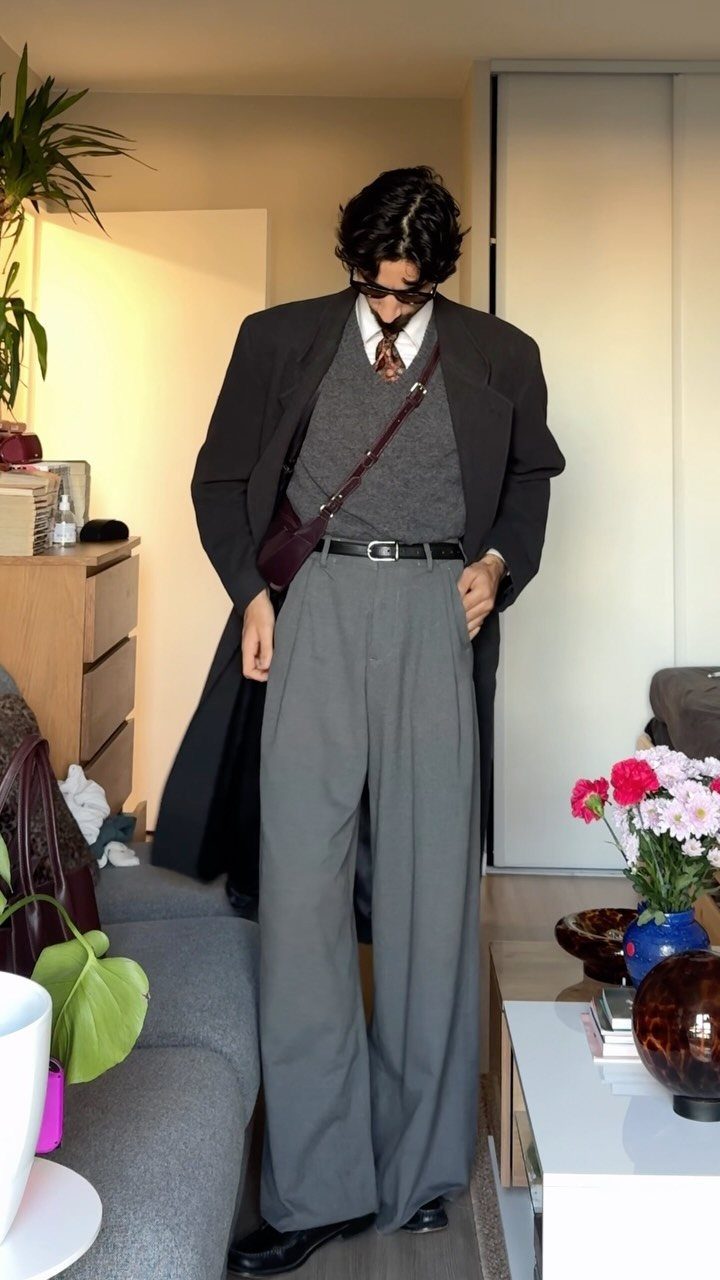 Man wearing a long black coat and wide-leg grey trousers in a living room.
