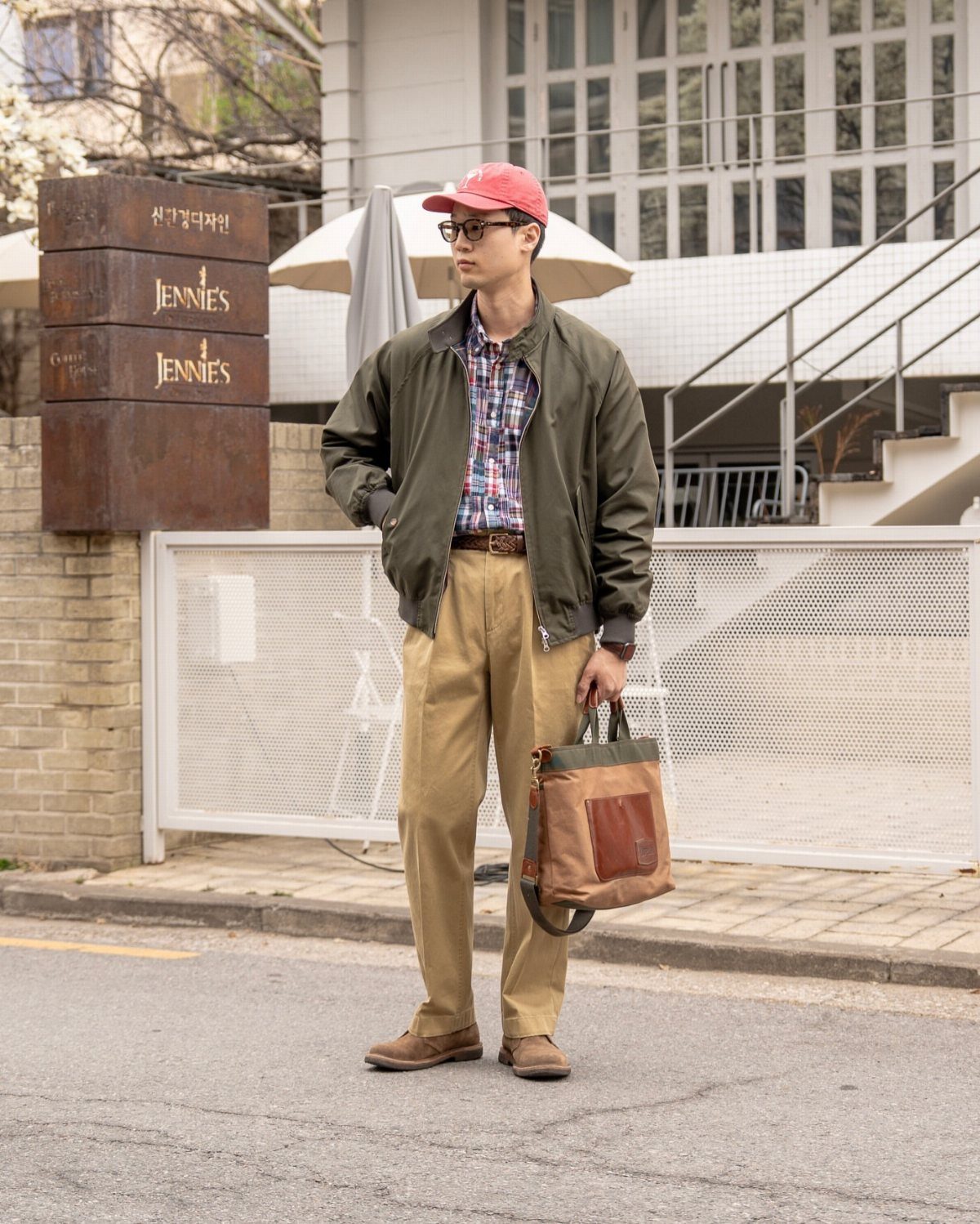 Man in red cap, olive bomber jacket, and khaki trousers holding a leather bag.