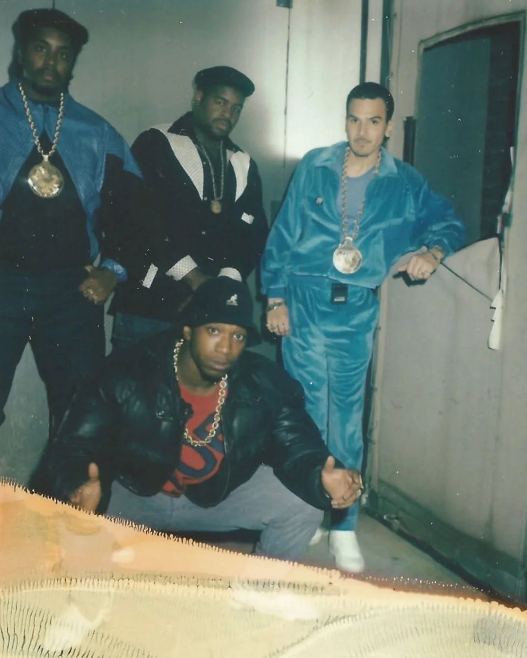 Four men wearing oversized jackets, gold chains, and tracksuits in 1980s hip-hop style.