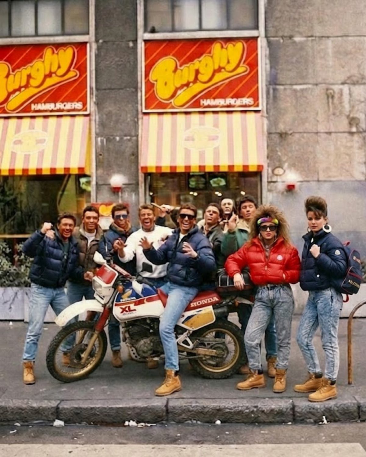 Group of men in 1980s puffer jackets and denim jeans posing by a motorcycle.
