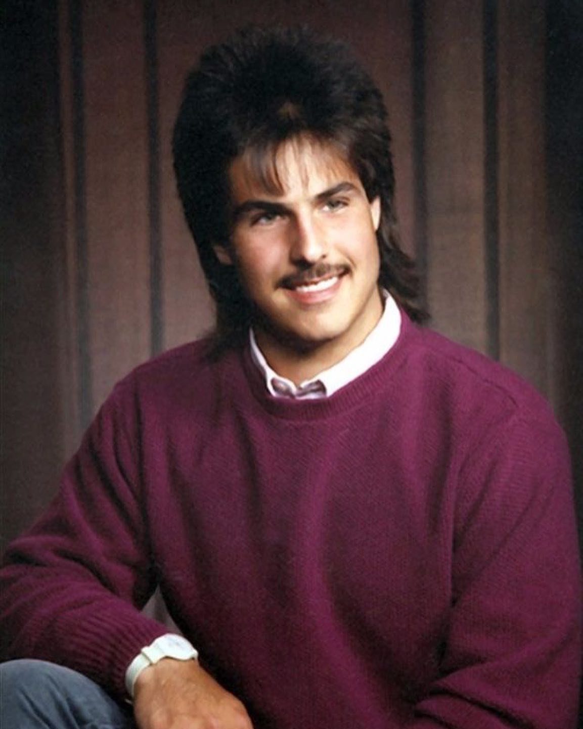 Man with mullet and mustache wearing a purple sweater and white collared shirt.