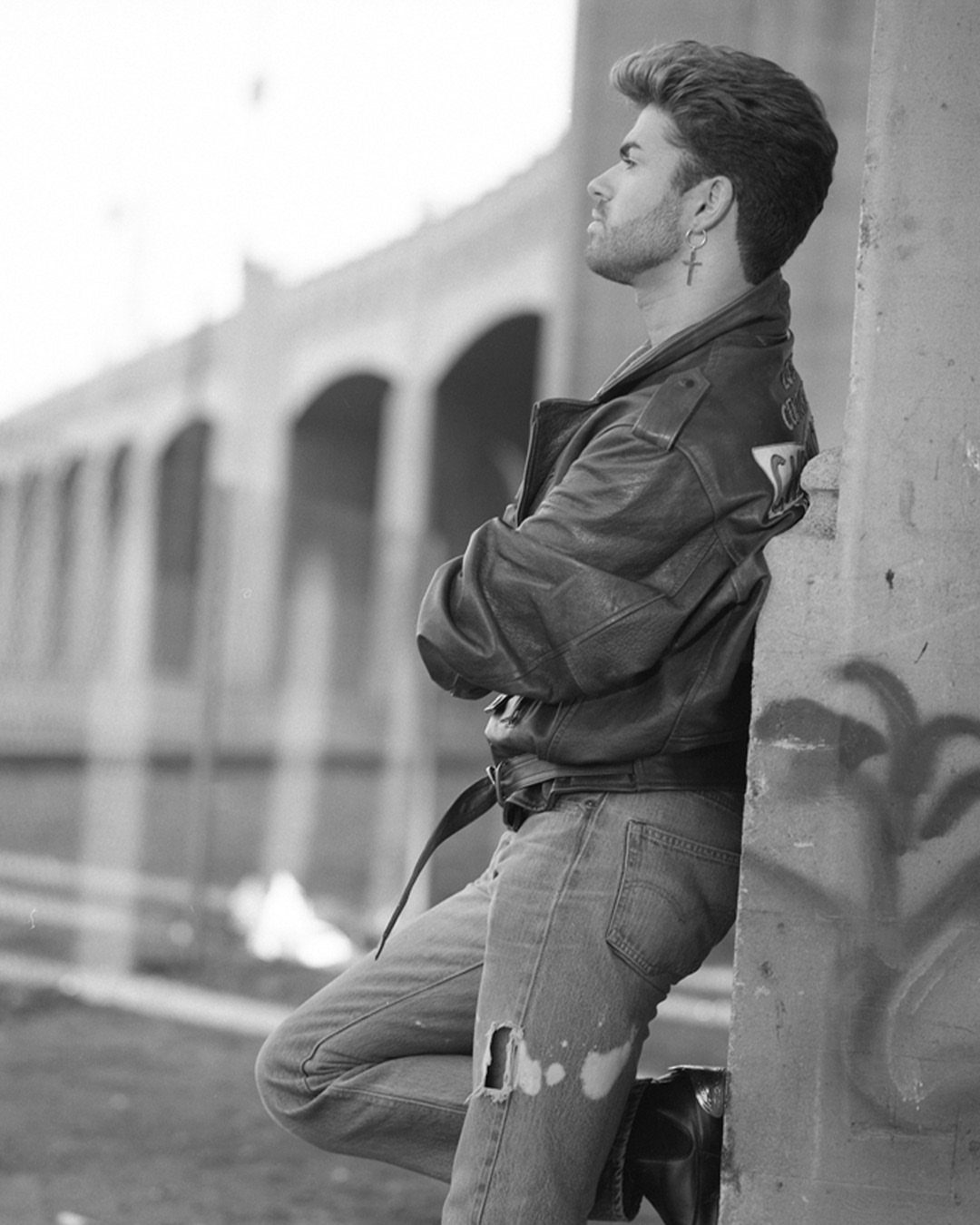 Man in leather jacket and ripped jeans leaning against a concrete wall.