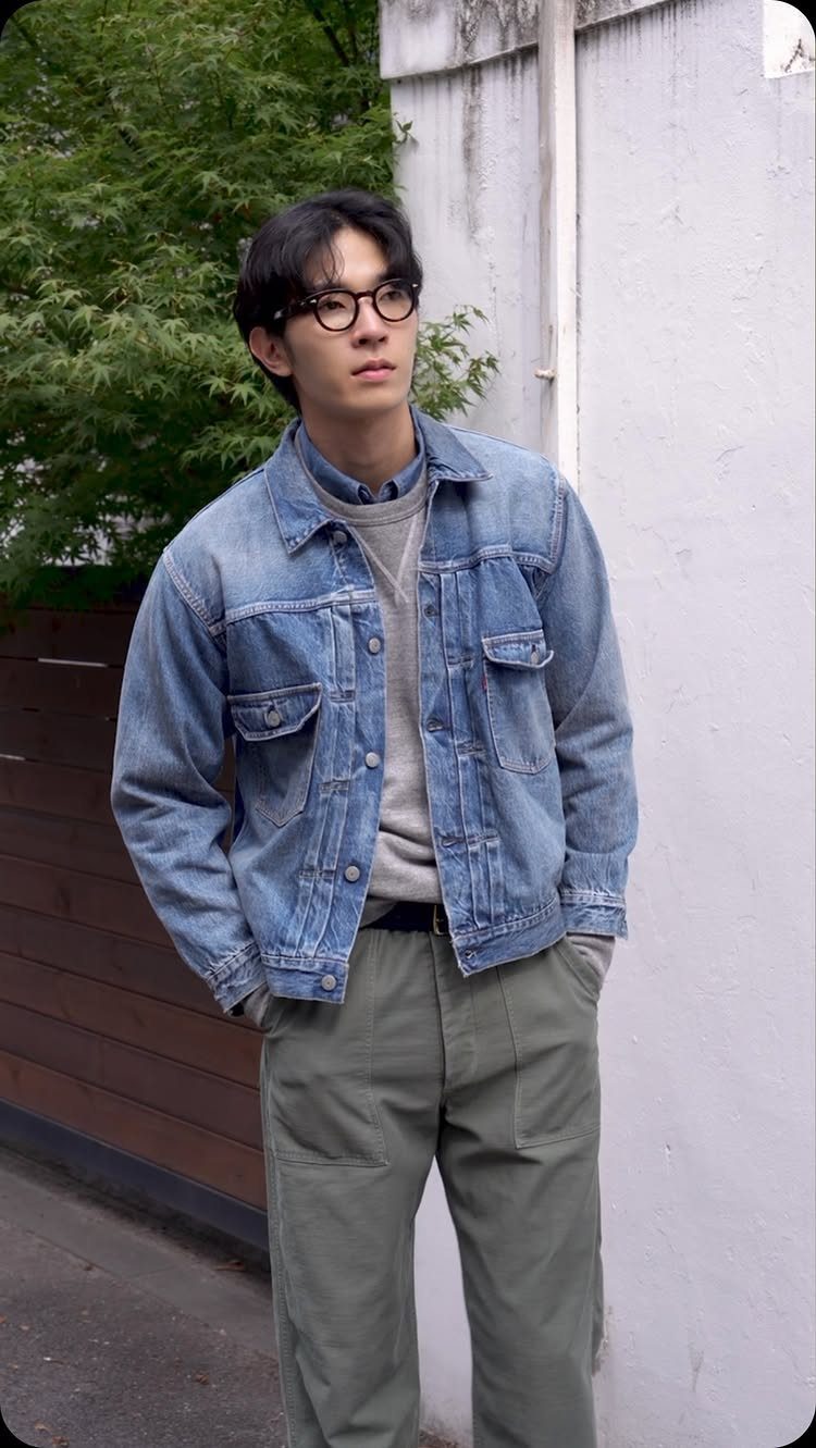 Man wearing a light blue denim jacket, grey sweater, and olive green trousers.