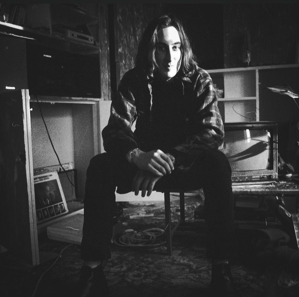 Man with long hair wearing a plaid flannel shirt sitting in a cluttered room.