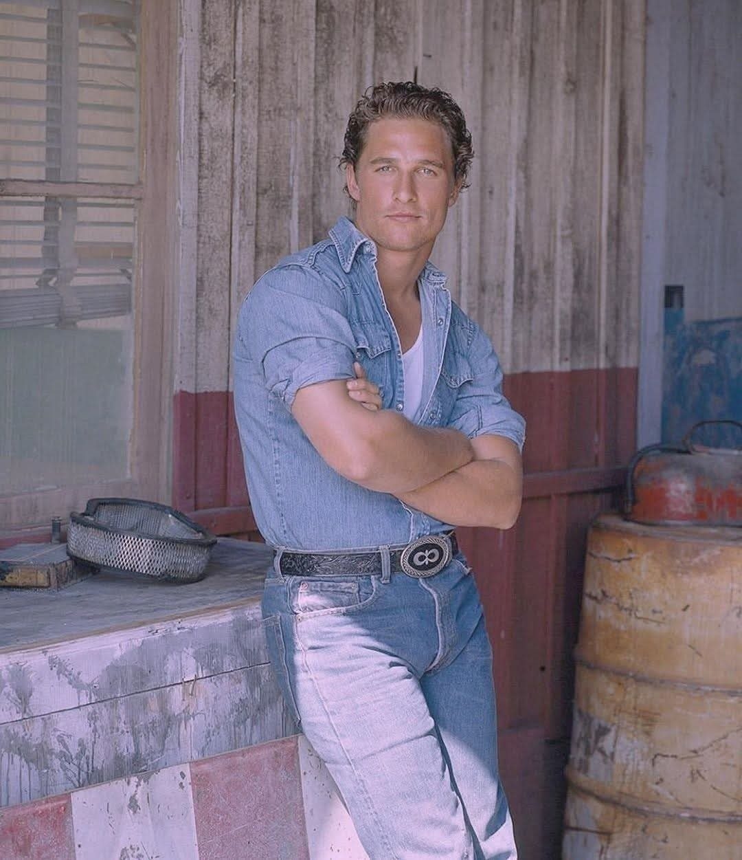 Man wearing a denim shirt and jeans leaning against a rustic wooden wall.