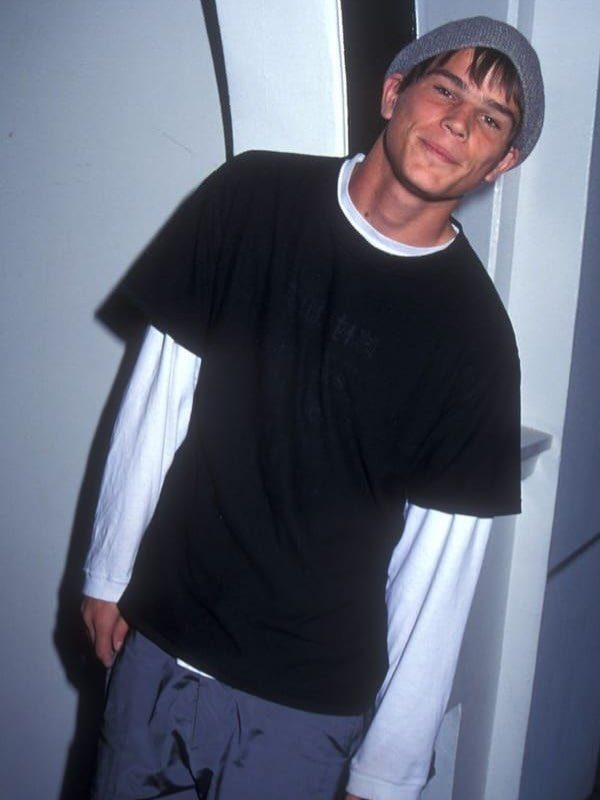 Young man wearing a beanie, layered t-shirts, and nylon pants in 90s style.