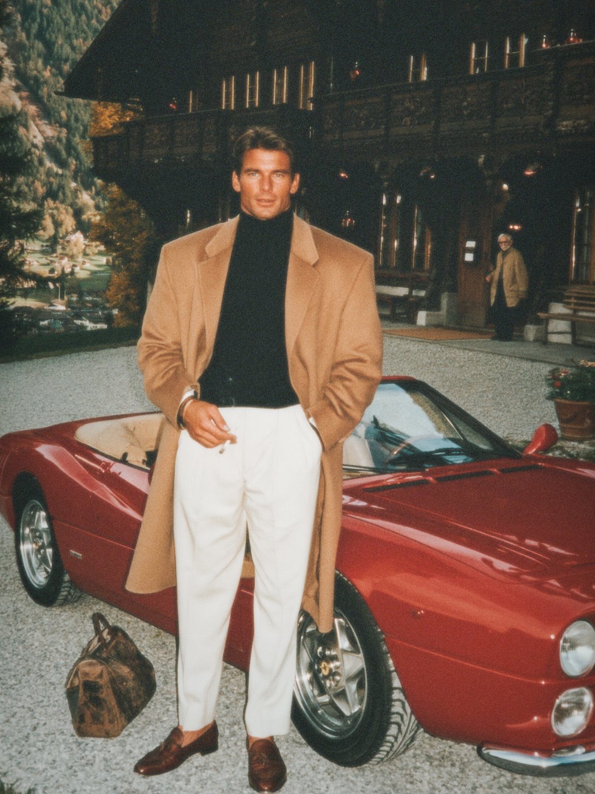 Man in black turtleneck and tan coat posing by red vintage convertible car.