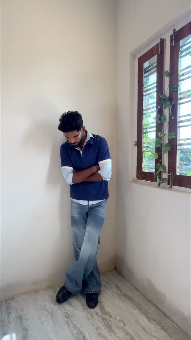 Man wearing a layered blue shirt and baggy jeans leaning against a wall.