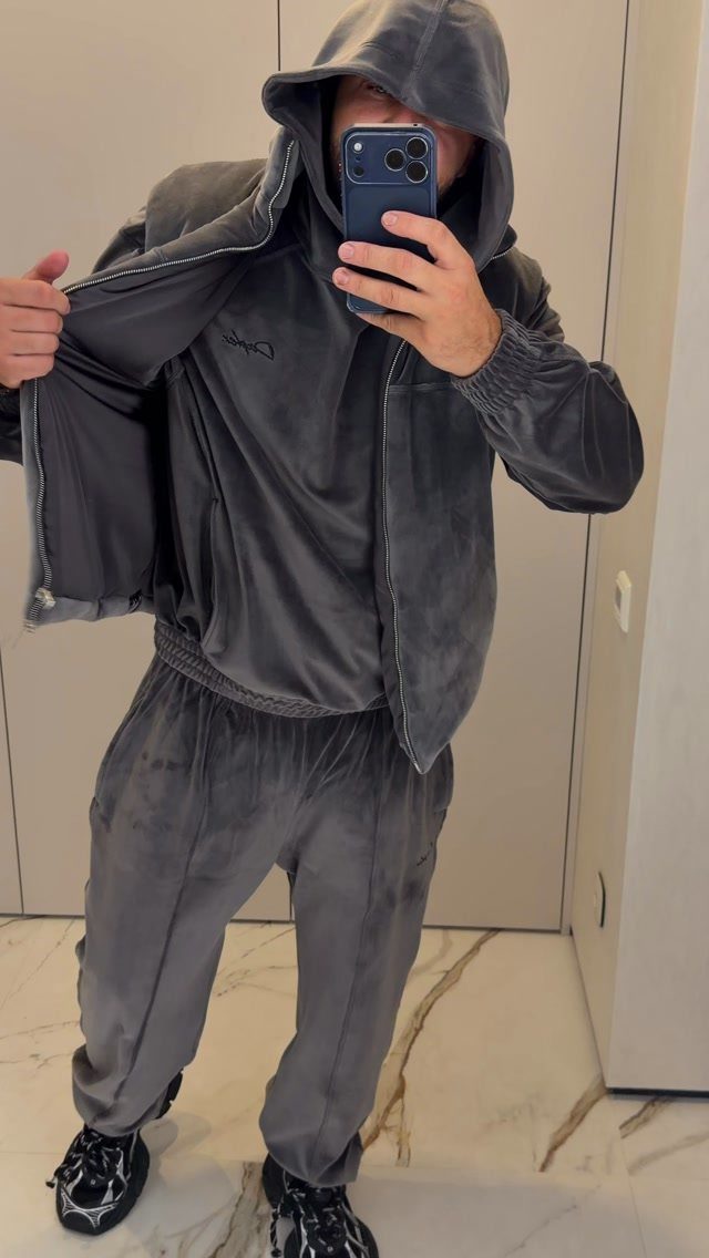 Man in a grey velour tracksuit taking a mirror selfie in a room.