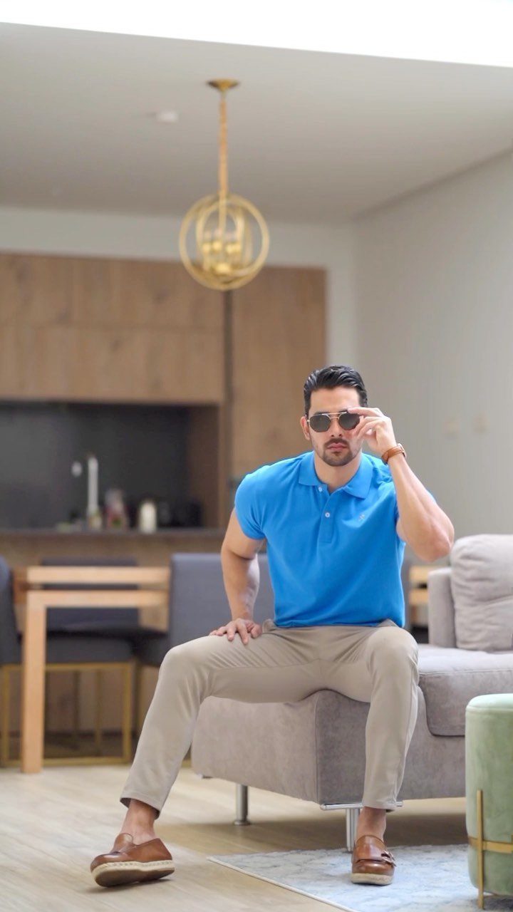 Man in blue polo and khaki pants sitting on a grey sofa indoors.