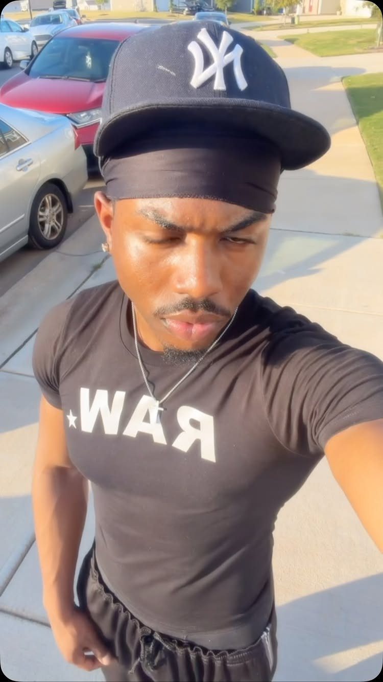Man in a black fitted RAW graphic t-shirt and NY baseball cap posing outdoors.