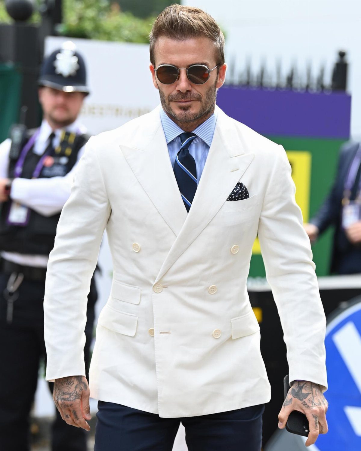 Man wearing a white double-breasted blazer, blue tie, and sunglasses outdoors.