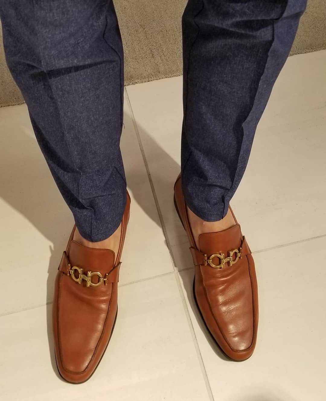 Brown leather horsebit loafers paired with blue trousers on a white tiled floor.