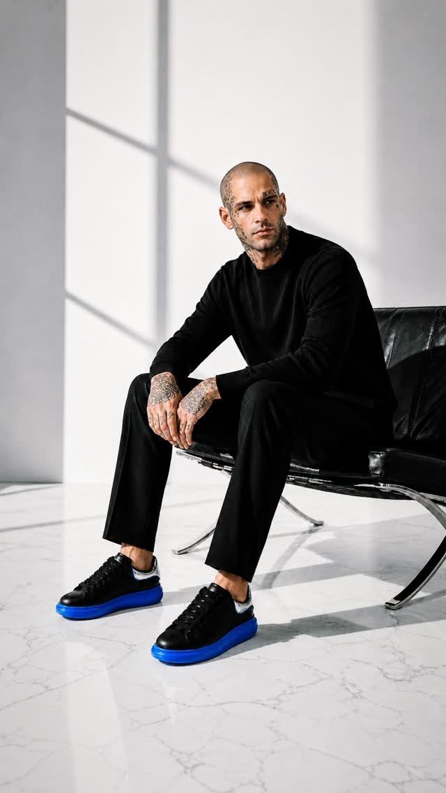 Man in black outfit wearing black sneakers with bright blue soles sitting on chair