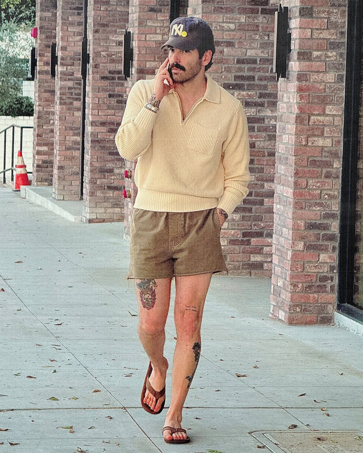Man wearing a cream polo and brown short shorts walking on a city sidewalk.