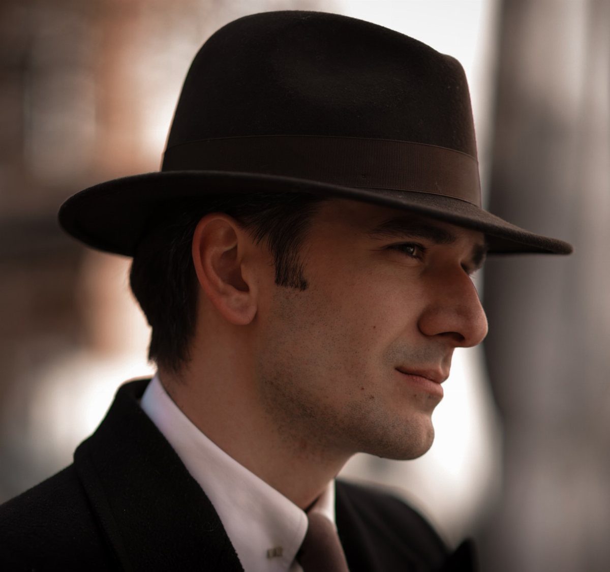 Man wearing a brown fedora, black suit, and white shirt in profile.