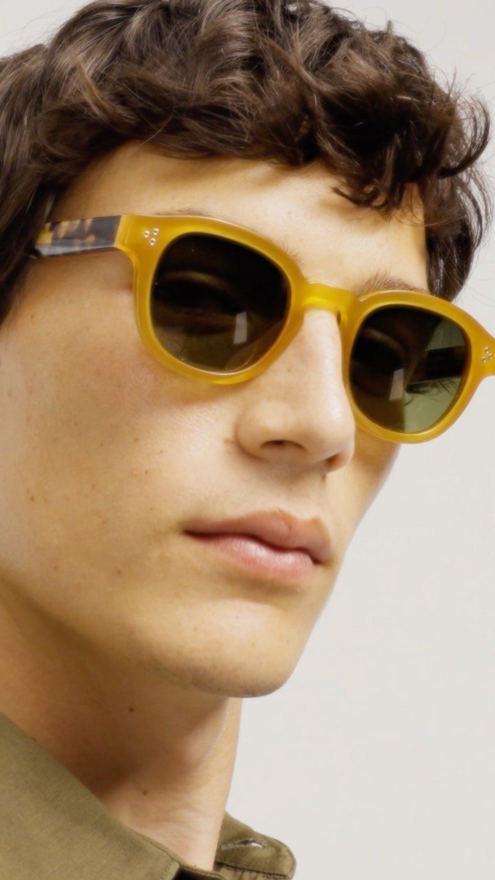 Young man with curly hair wearing bold yellow mod-style sunglasses and brown shirt.