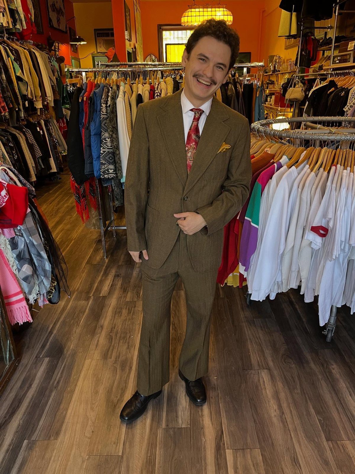 Man in a brown tailored suit and red tie standing in a vintage shop.