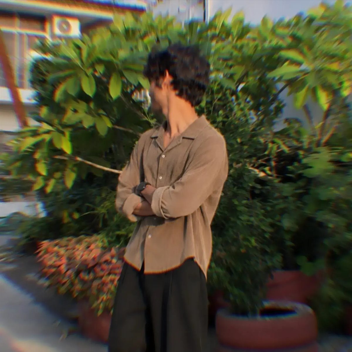 Man wearing a brown button-down shirt and black wide-leg trousers standing by green bushes.