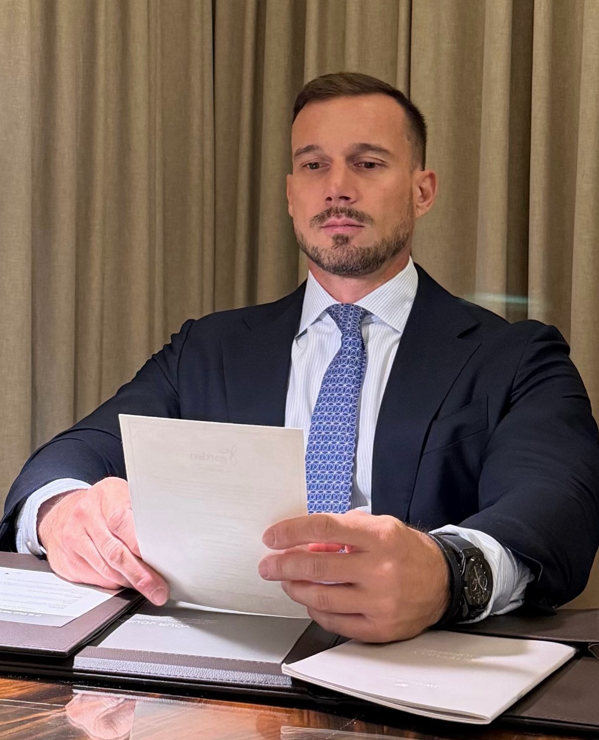 Man in navy blue suit and tie holding a document at a desk.
