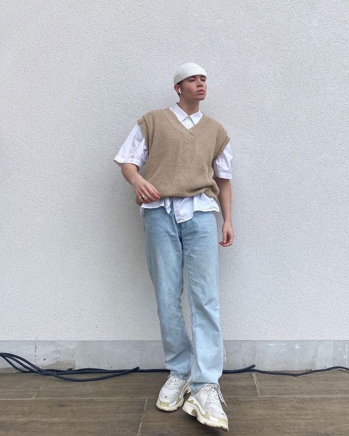 Man in tan knit vest, white shirt, light jeans and white beanie posing.