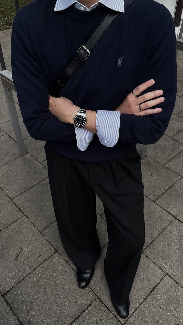 Man in navy sweater, striped shirt, and black trousers standing on grey pavement.