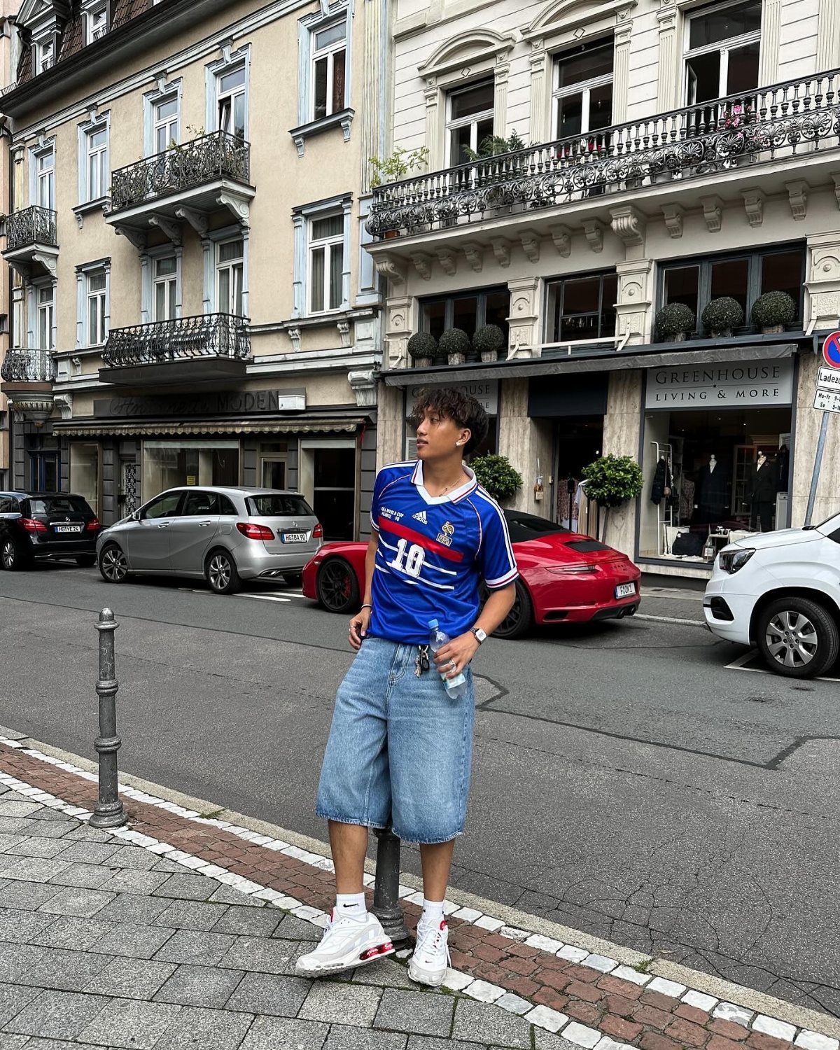 Man wearing a blue soccer jersey and loose denim shorts on a city street.