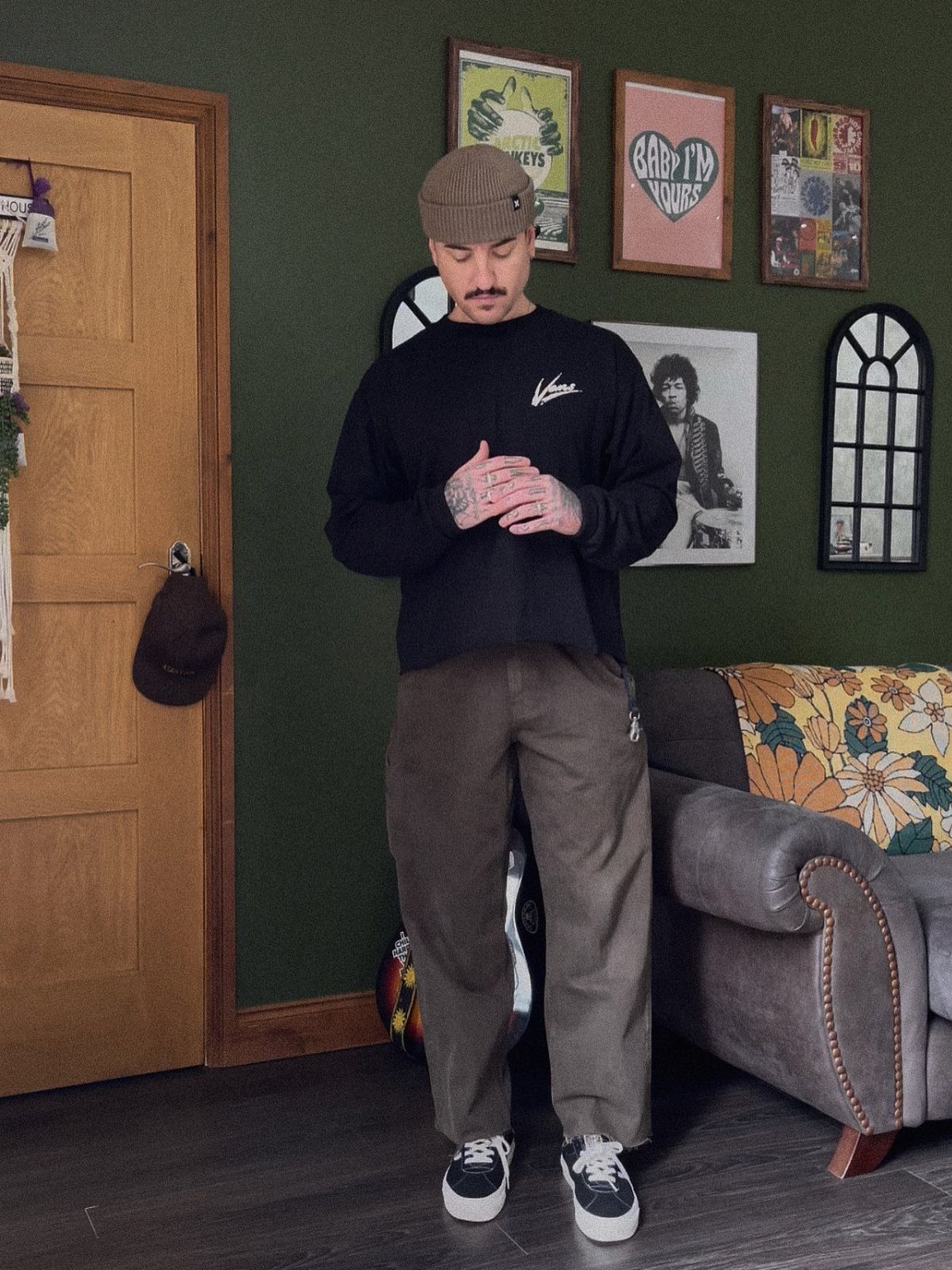 Man in brown beanie, black shirt, and loose brown trousers posing indoors.