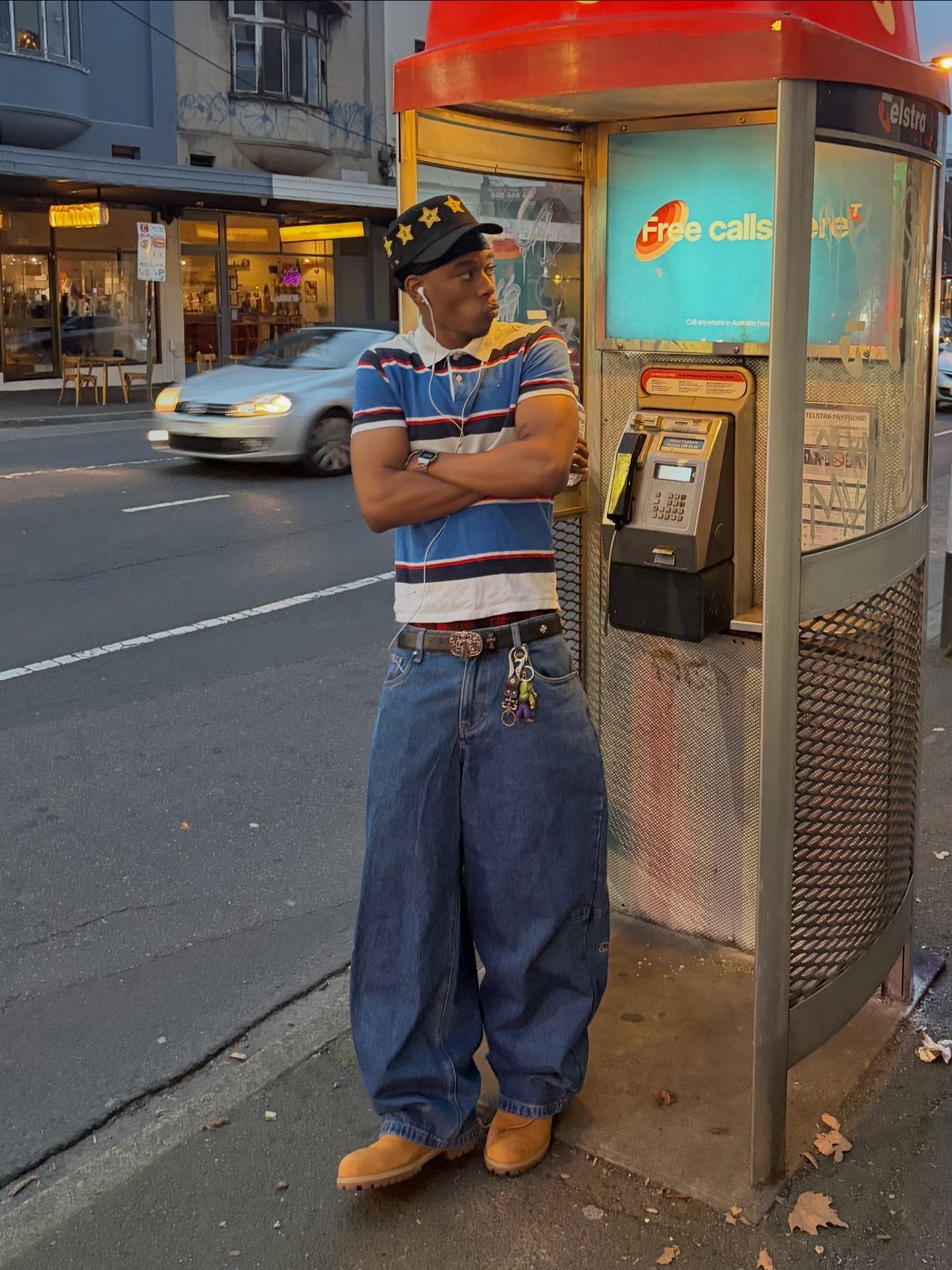 Man in baggy jeans and striped shirt posing by a red phone booth.