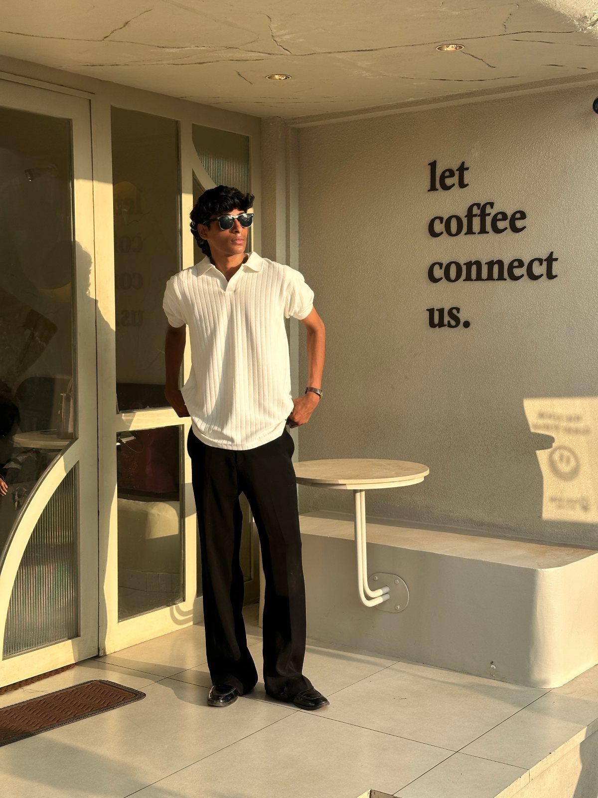 Man in white button-down shirt and wide-leg black trousers posing by a cafe wall.