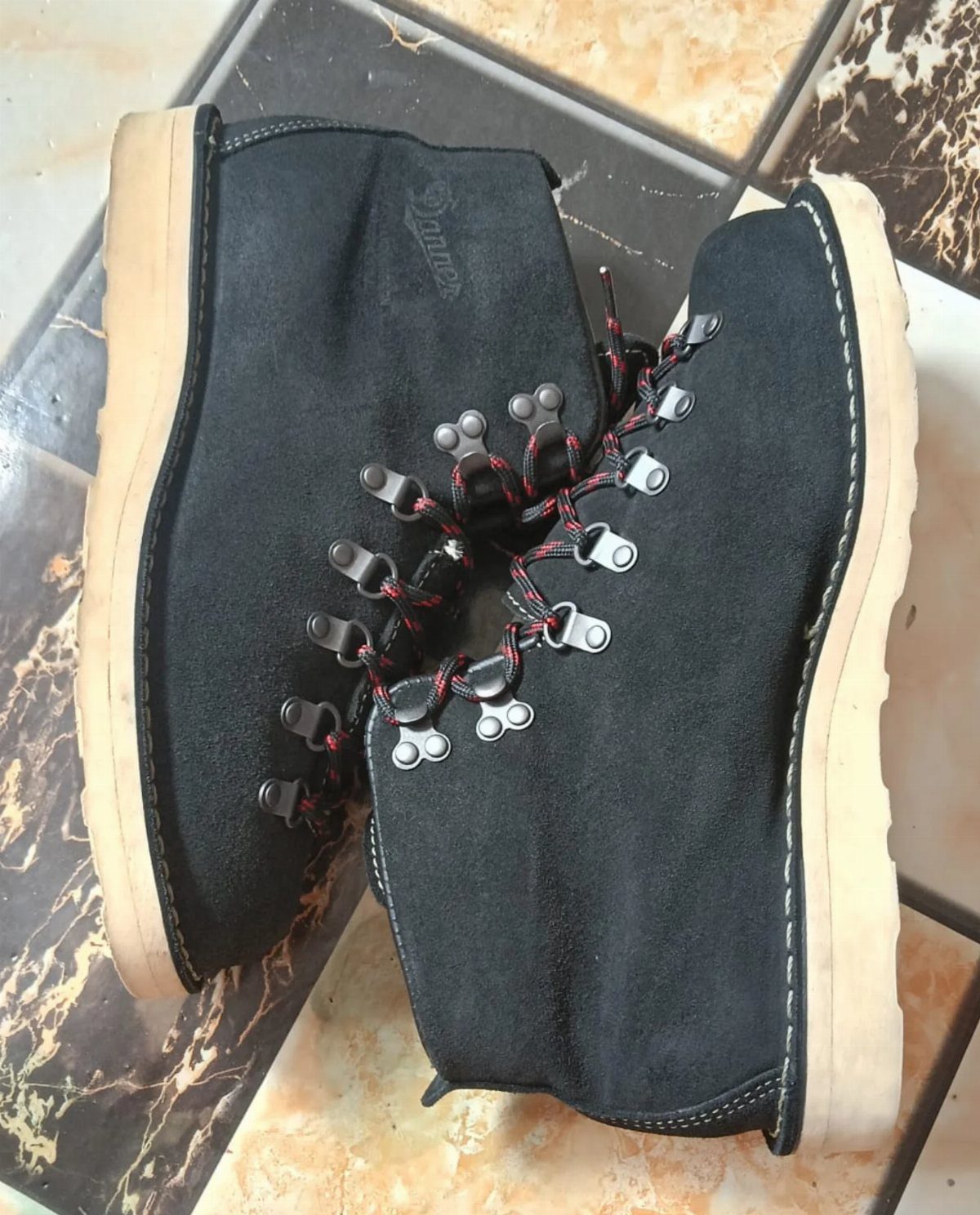 Black suede winter boots with red laces and tan soles on marbled tiles.