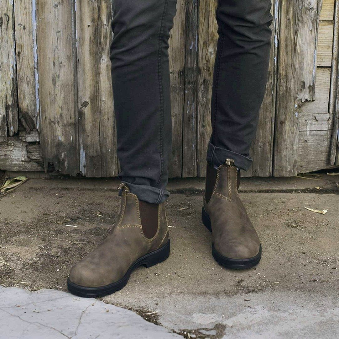 Brown leather Chelsea boots worn with dark grey trousers against a wooden fence.