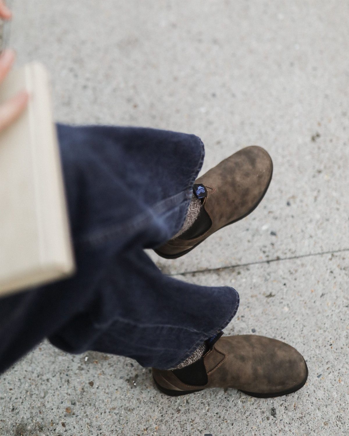 Brown suede ankle boots and dark blue trousers on a grey concrete ground.