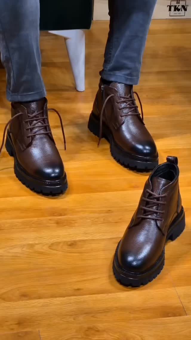 Dark brown leather lace-up winter boots worn with grey trousers on wooden flooring